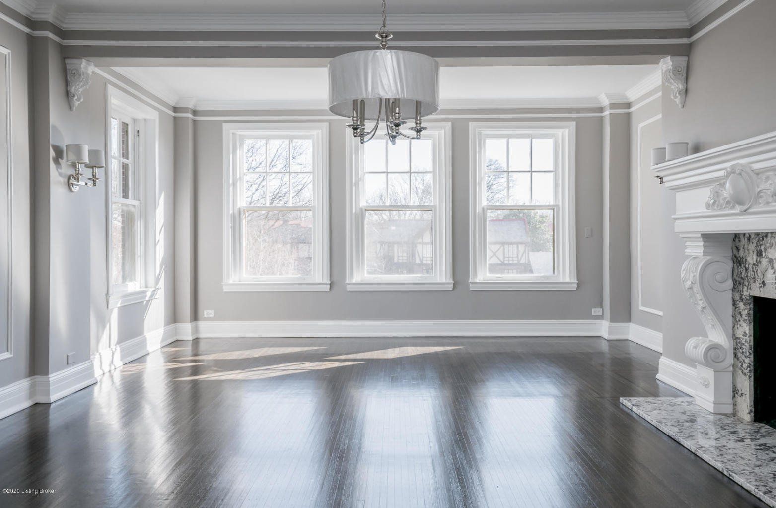 Empty living room with three windows, a fireplace, and a chandelier. Dark wood floor.