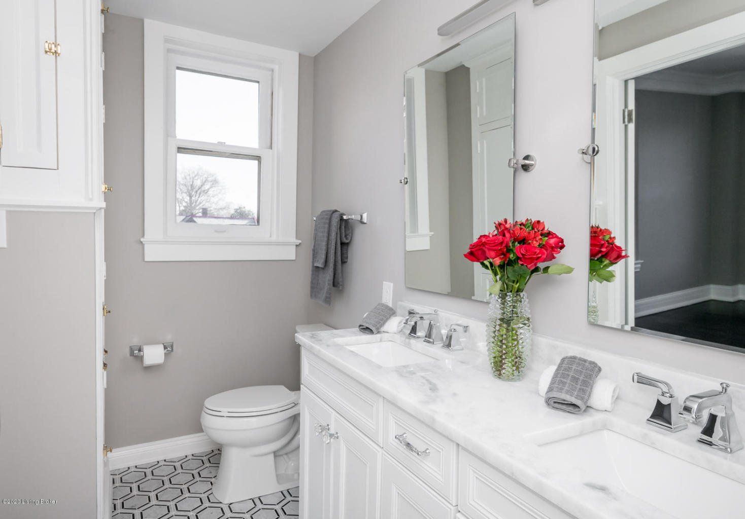 Bright bathroom with white vanity, mirror, toilet, patterned floor, and red flowers in a vase.