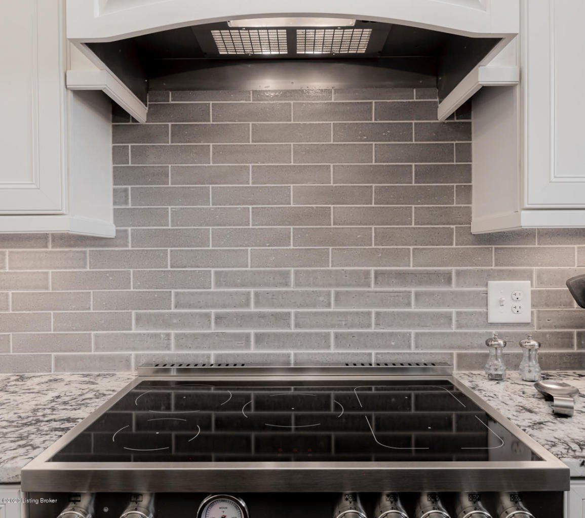 Kitchen with grey brick backsplash, cooktop, and range hood. Cabinets frame the view.