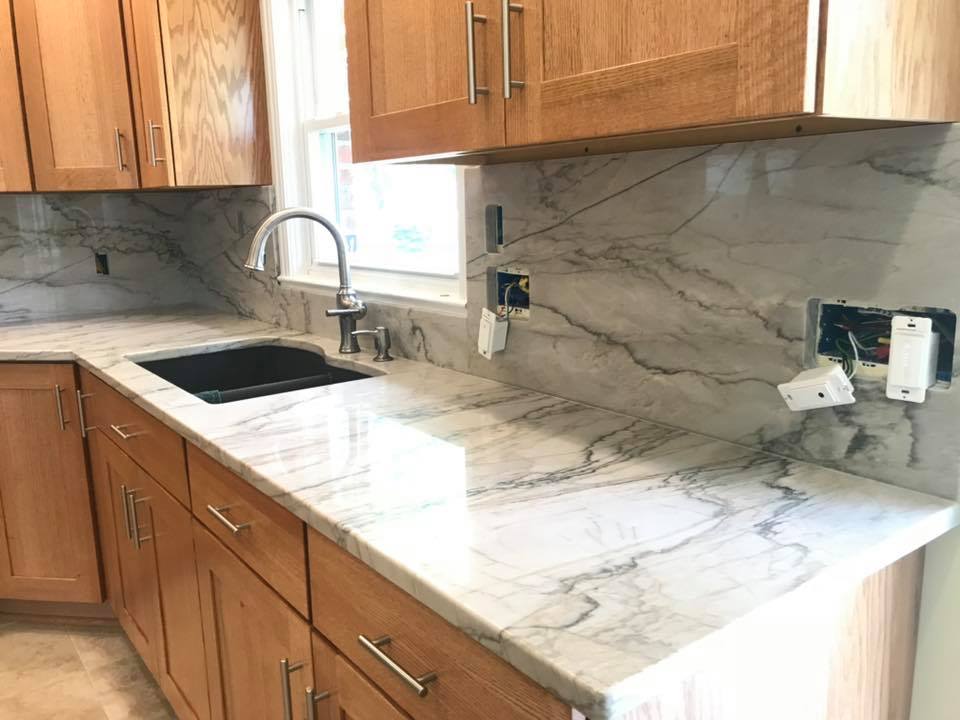 Modern kitchen with wood cabinets, marble countertops, sink, and wall outlets under a marble backsplash