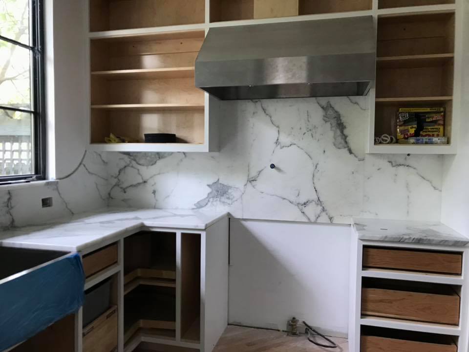 Kitchen remodel with marble backsplash and countertops, stainless steel hood, and unfinished cabinets and shelves.