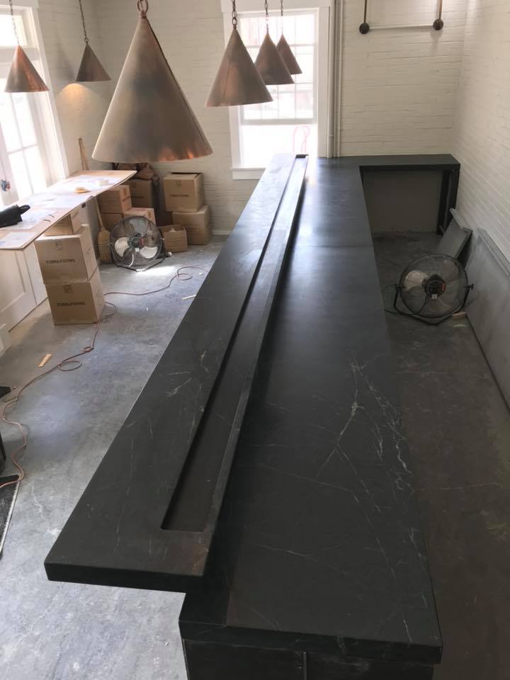 Long, dark countertop with inset, under construction, near a window. Copper pendant lights hang above.