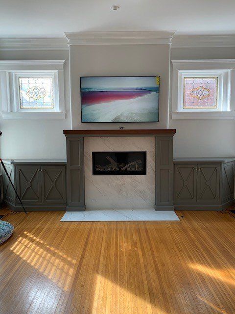 Living room with fireplace, TV, wood floors, and two small windows with art. Grey and wood tones dominate.