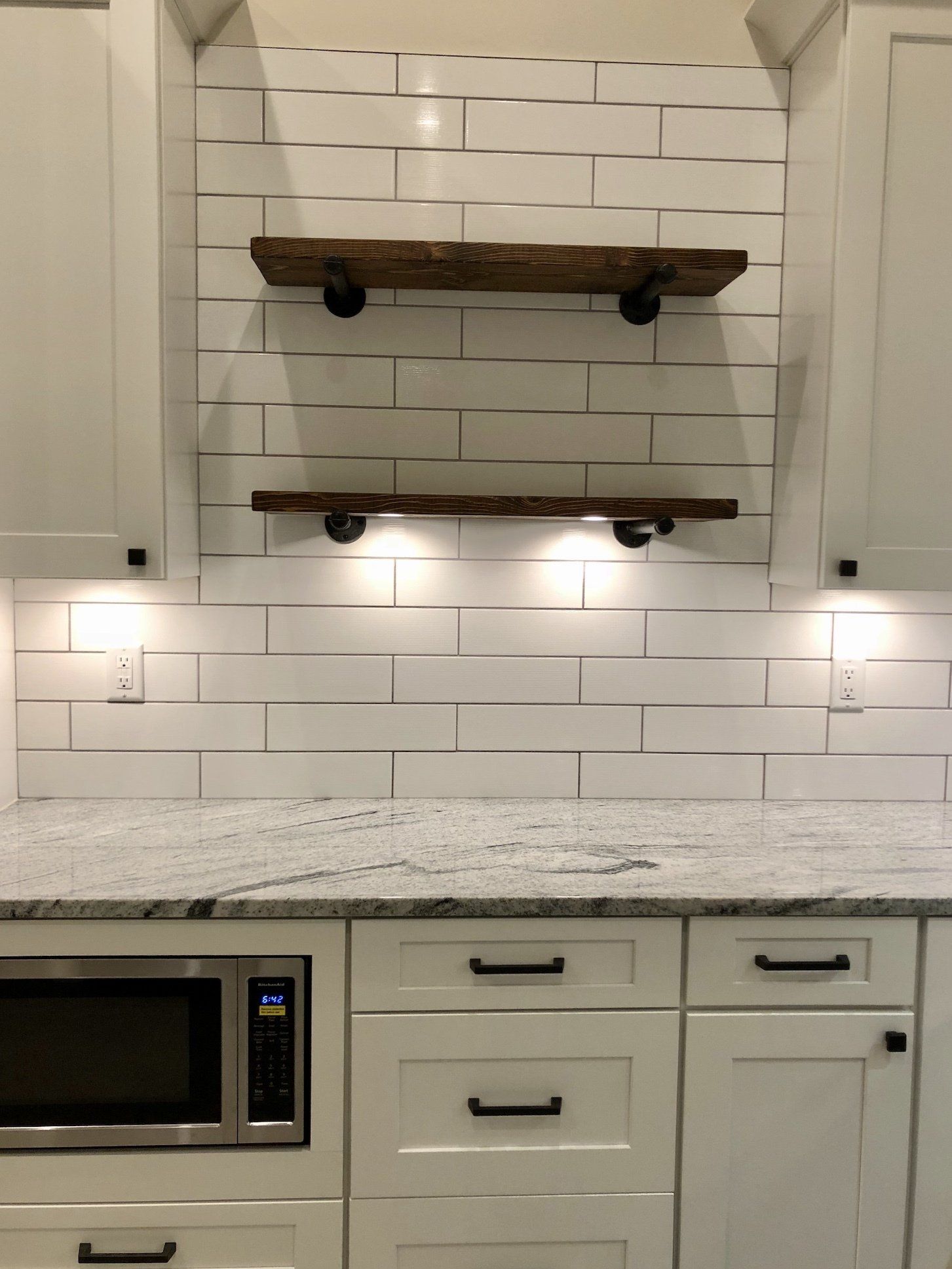 White kitchen with granite countertop, white subway tile backsplash, and two wooden shelves.