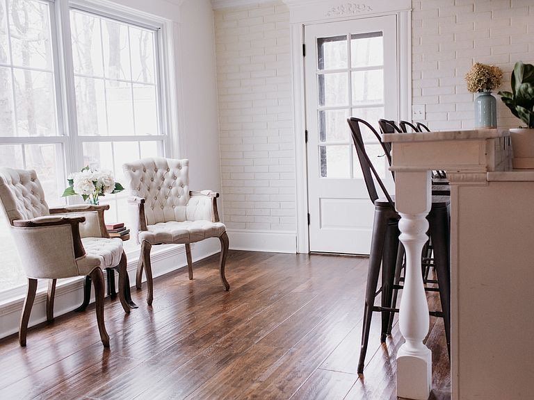 Elegant dining area with tufted chairs by window, brick wall, dark wood floors, and white door.