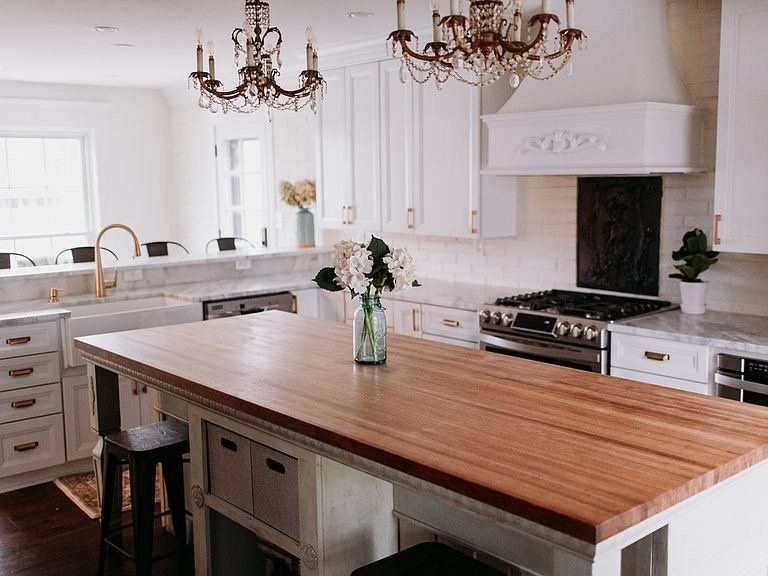 Elegant white kitchen with wooden island, gold hardware, and two chandeliers.