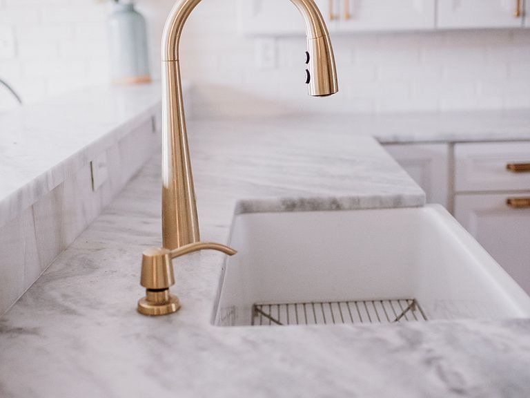 Gold faucet over a white farmhouse sink on a marble countertop in a kitchen.