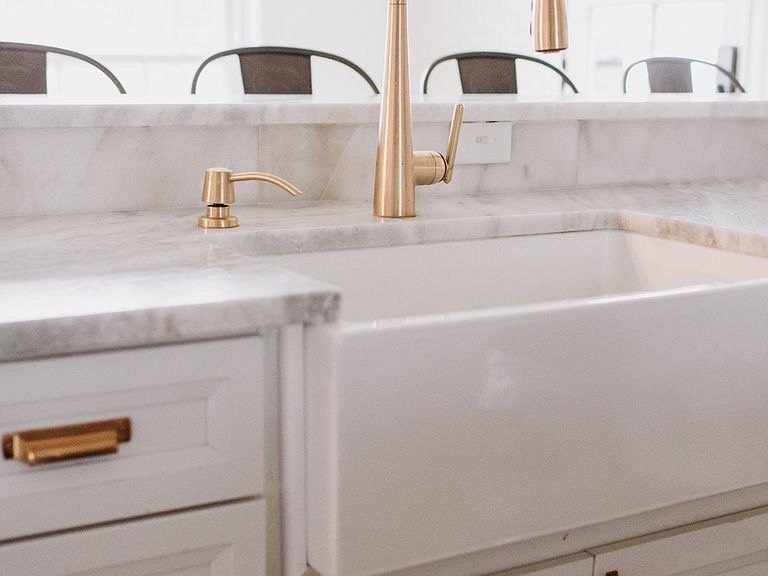 White farmhouse sink with gold faucet and soap dispenser on a marble countertop. White cabinets below.