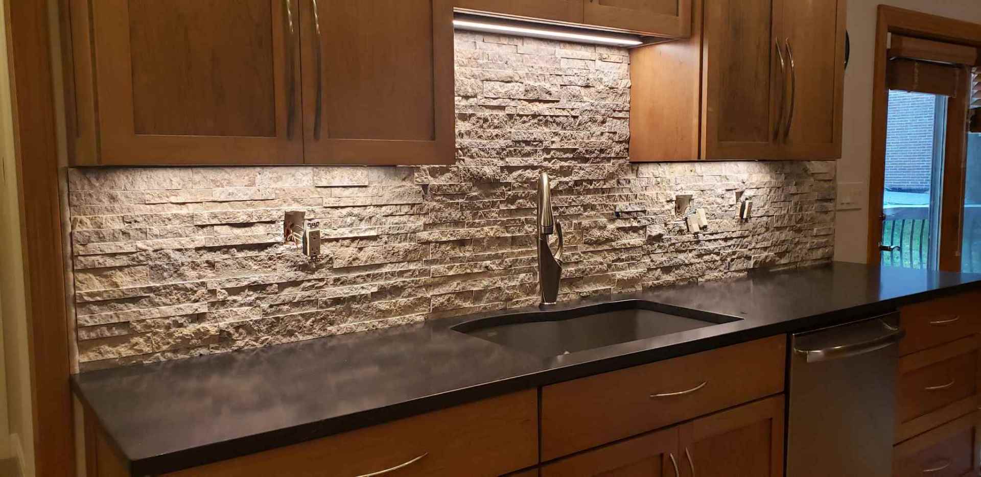 Kitchen with stone backsplash, dark countertop, wooden cabinets, and a stainless steel sink and faucet.