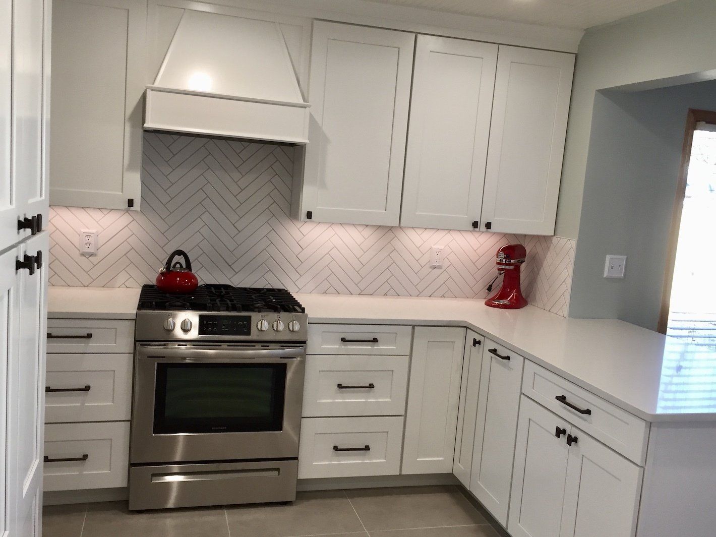 White kitchen with stainless steel appliances, herringbone backsplash, and red accents.