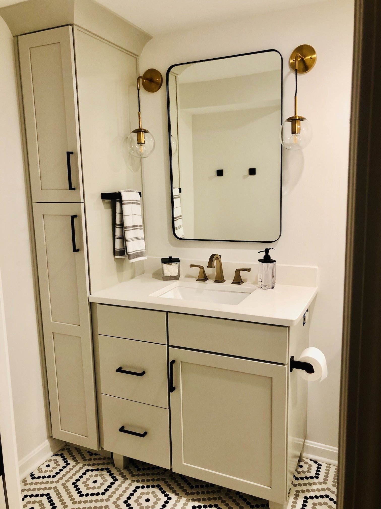 Beige bathroom vanity with mirror, brass sconces, and patterned black-and-white floor tiles.