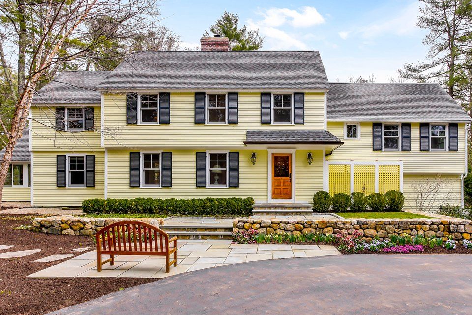 Two-story yellow suburban house with brown shutters, front porch, and landscaped garden.