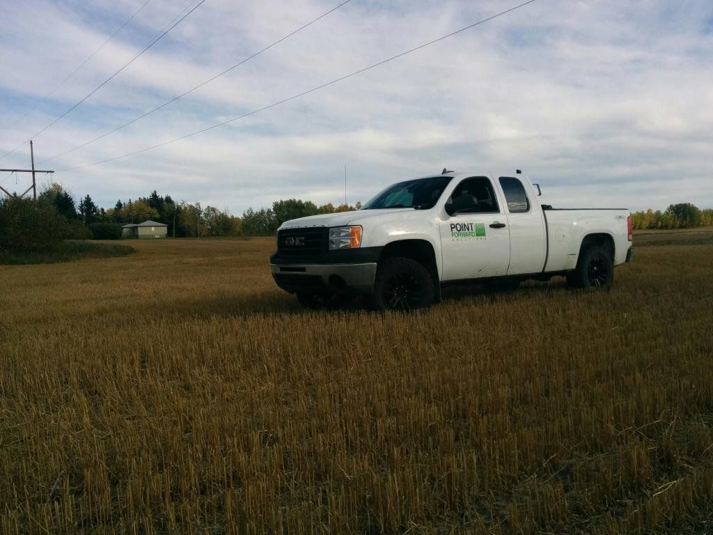 A white branded point forward solutions GMC pickup truck parked in a large, dry harvested field under a cloudy sky.