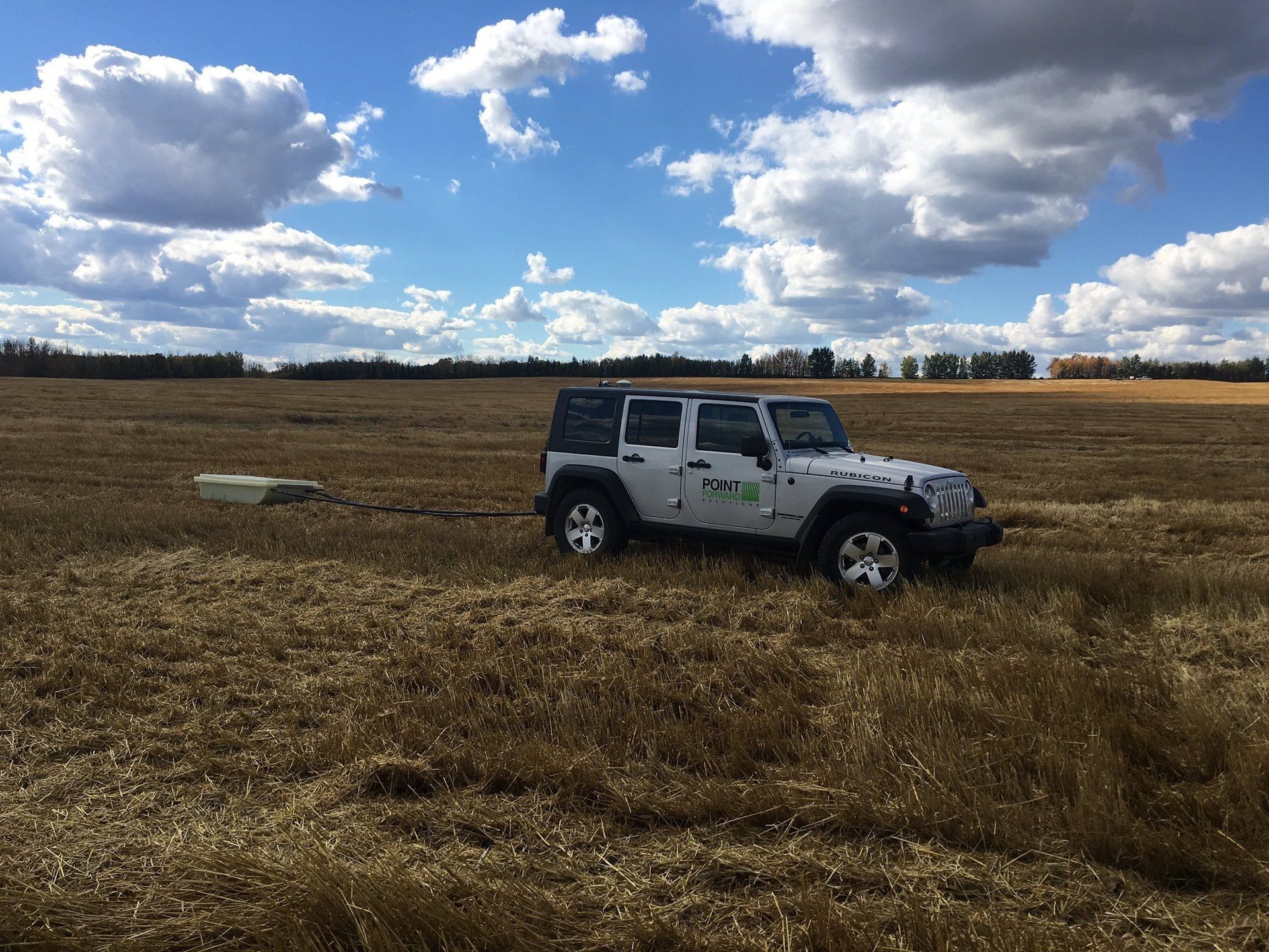 A white Jeep is parked in a large, harvested field under a partly cloudy blue sky doing EM38 field mapping