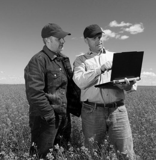 Two people wearing caps stand in a field, looking at a laptop held by one person pointing at the screen.