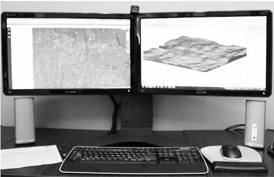 A desk setup with two monitors showing geographic mapping software, a keyboard, mouse, and speakers on a flat surface.