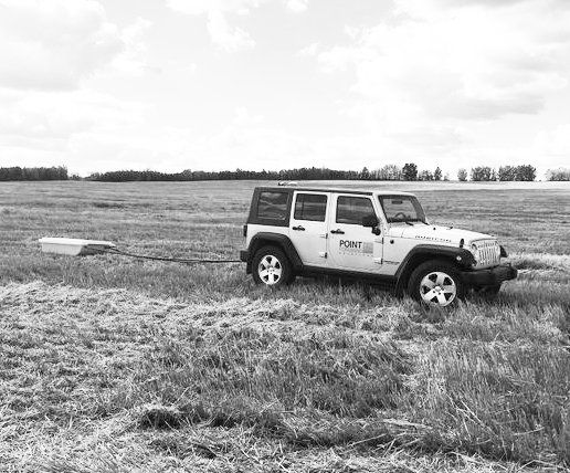 A white Jeep towing a ground-penetrating radar sled across a grassy field under a cloudy sky.