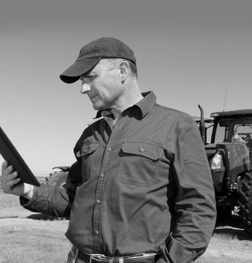 A person in a cap and collared shirt stands in a field holding a tablet, with a tractor visible in the background.