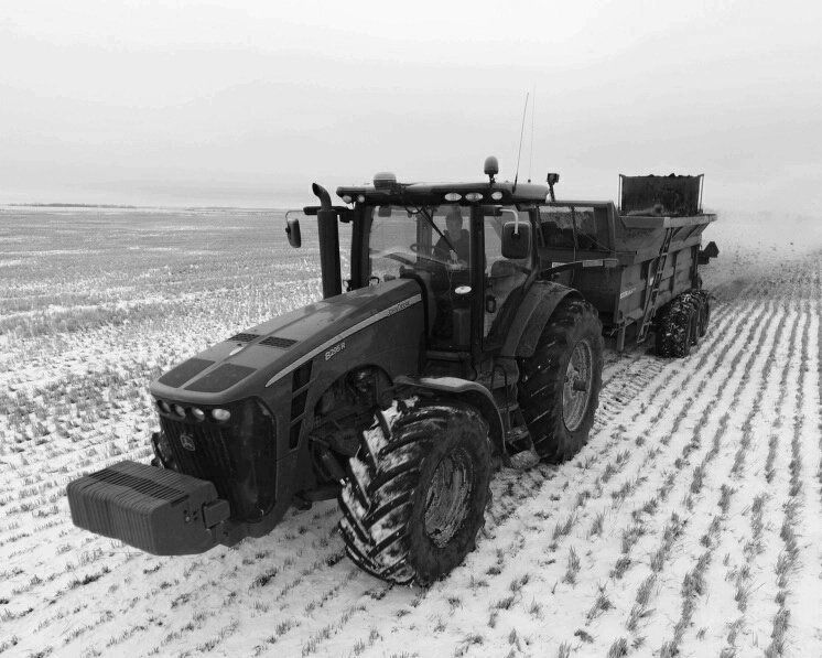 A tractor pulling a spreader through a snow-covered field, performing agricultural work in a bleak, wintry landscape.