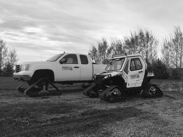 A white truck and a white UTV, both equipped with snow tracks, parked on a dirt lot with trees in the background.