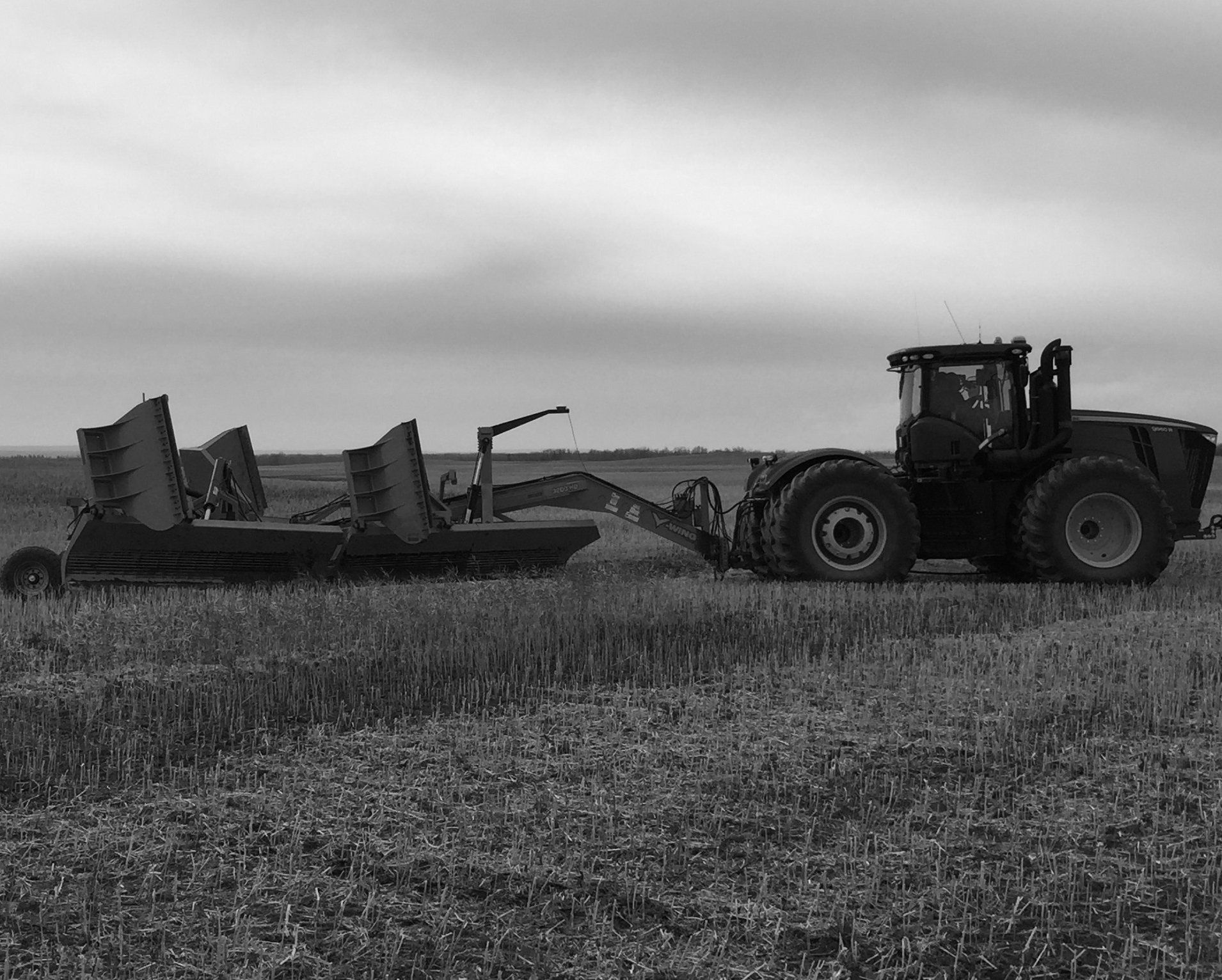 A large tractor pulls an agricultural scraper implement across a harvested field under a cloudy sky.