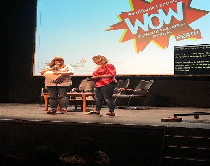 Women presenting on stage with captions on plasma screen to  the side.