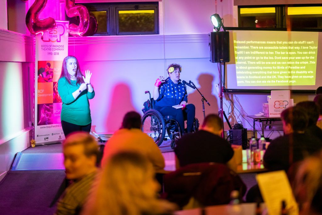 Performer on wheelchair on stage with BSL interpreter and captioned screen. Performer on wheelchair on stage with BSL interpreter and captioned screen.