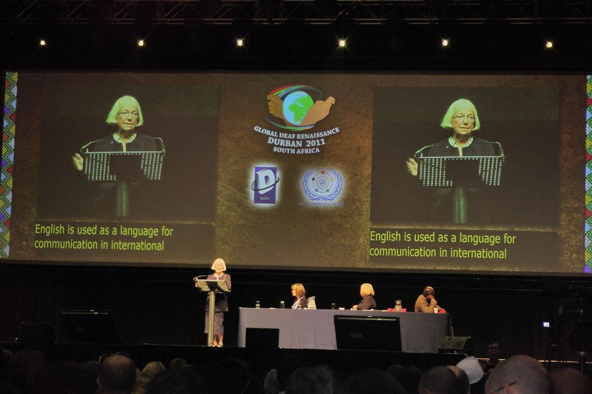 Woman at lecturn in front of large projection with overlaid captions.