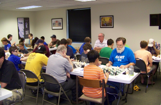 A group of people are playing chess and one man is wearing a shirt that says gems