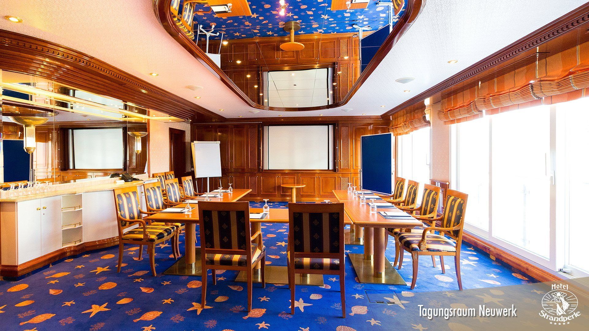 A conference room with tables and chairs and a large screen on the ceiling.