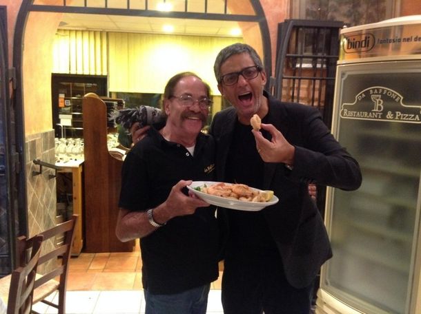Due persone in posa con un piatto di cibo all'ingresso di un ristorante, sorridenti e divertite.