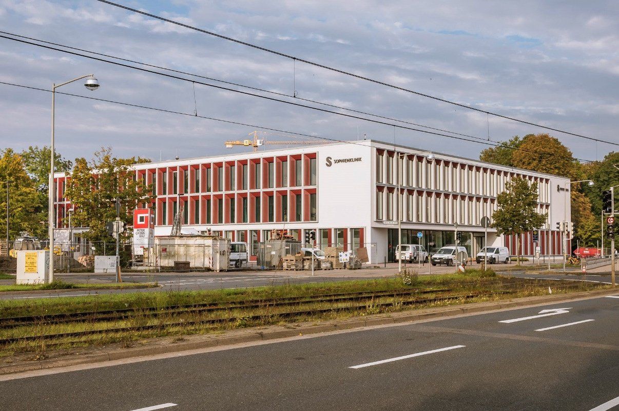 Modernes weißes Gebäude mit roten Fensterrahmen; Straße und Straßenbahnschienen im Vordergrund.