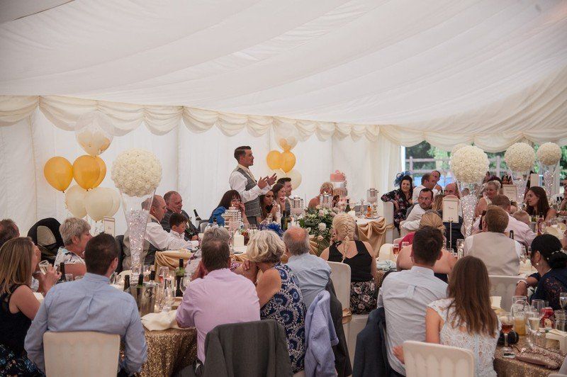 A man is giving a speech at a wedding reception in a tent.