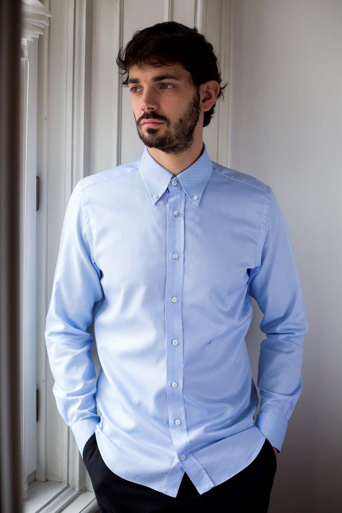 Un uomo con una camicia azzurra abbottonata è in piedi in un ambiente interno, vicino a una finestra, e guarda di lato.