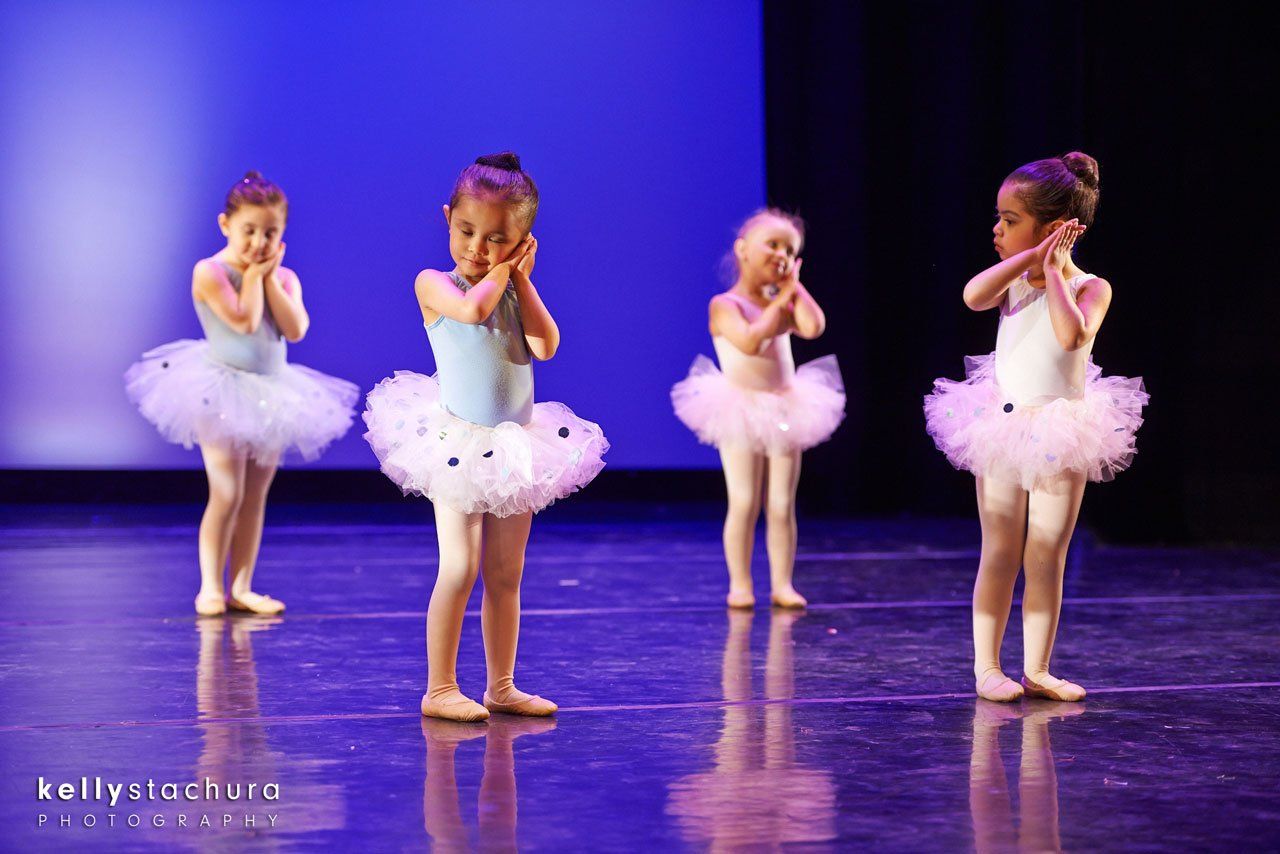 Four young ballet dancers in white tutus pose on a purple-lit stage.