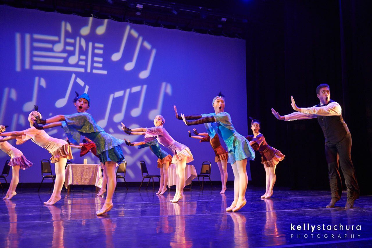 Ballet dancers in colorful costumes perform on a lit stage with musical note backdrop.