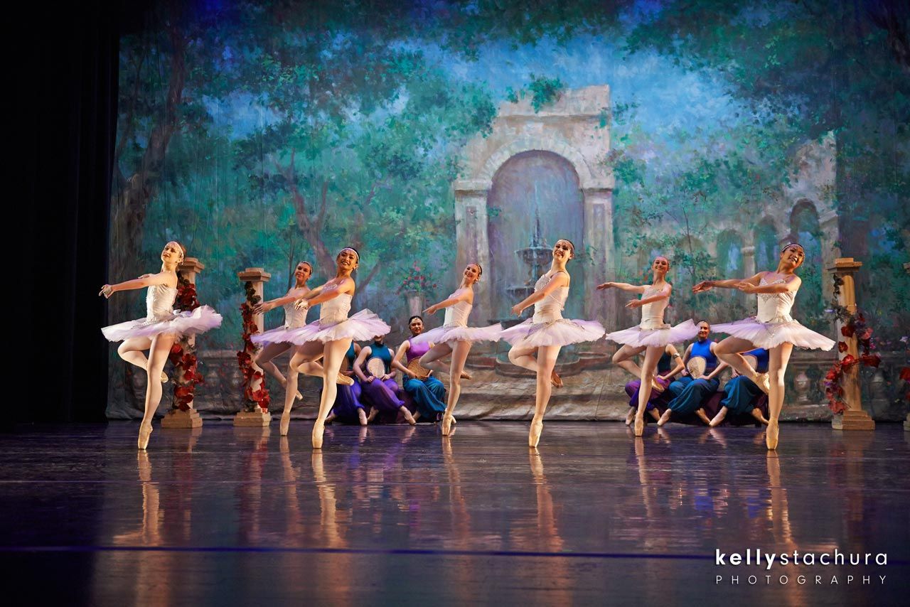 Ballet dancers in white tutus perform onstage before a painted garden arch backdrop.