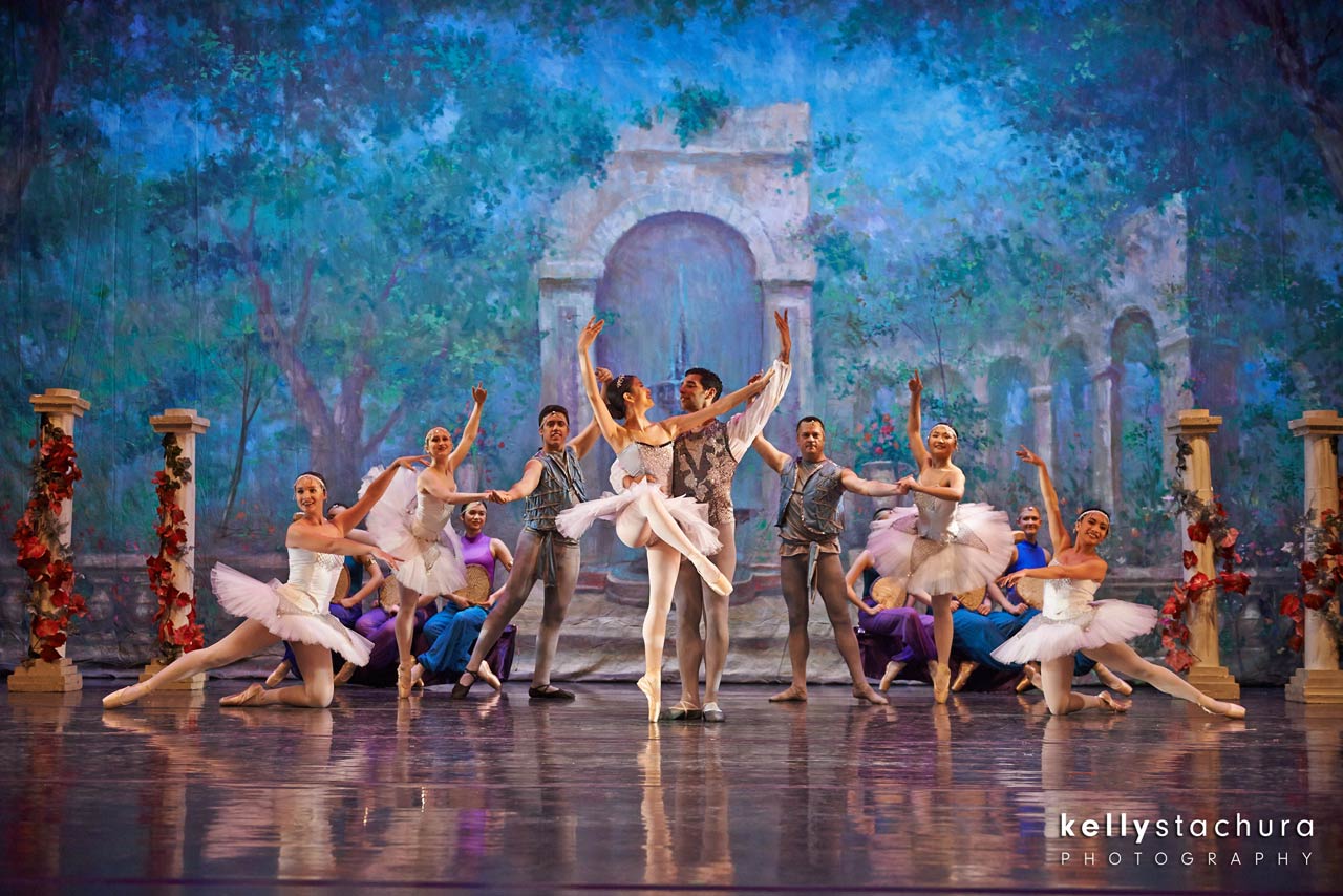 Ballet troupe in white costumes poses onstage before a blue-lit arch with ornate columns and painted backdrop