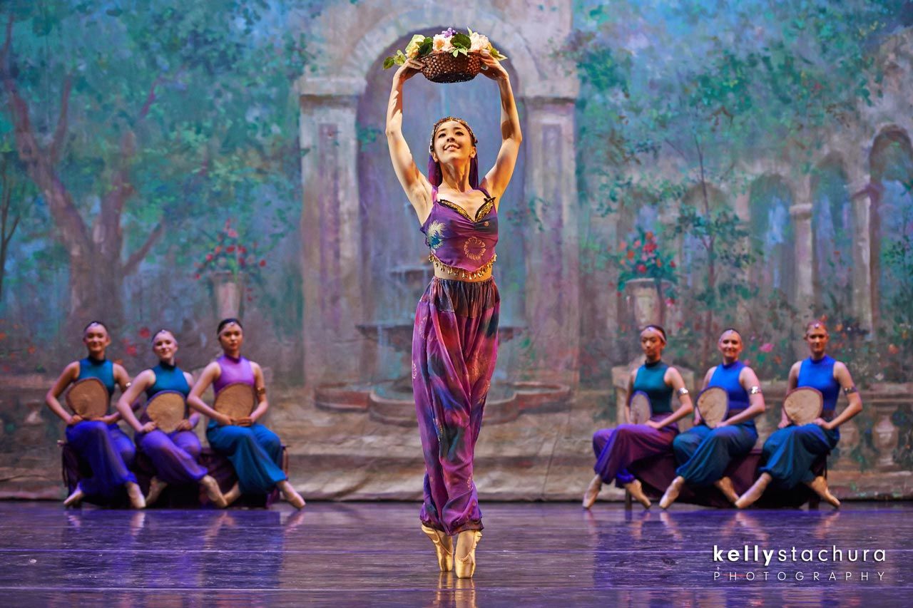 Ballet dancer in purple costume balancing a floral basket overhead on stage, with seated dancers behind her