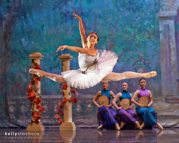Ballet dancer in white tutu performs a leap onstage, with seated dancers and blue-purple backdrop behind.