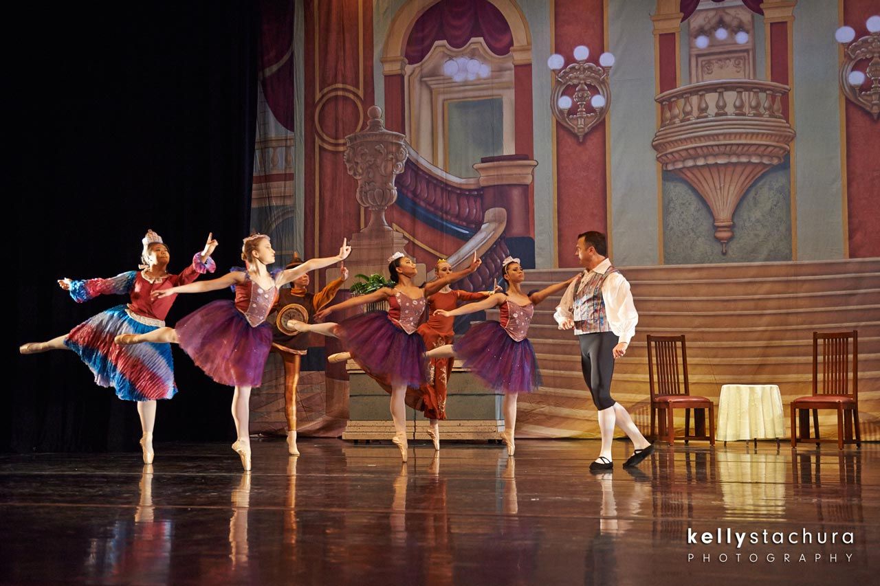 Ballet dancers in colorful costumes perform on a theater stage with a painted palace backdrop