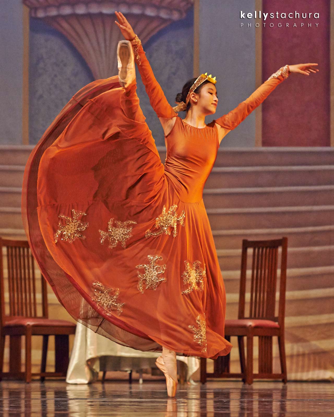 Orange-clad dancer performing a graceful ballet pose onstage with chairs in the background