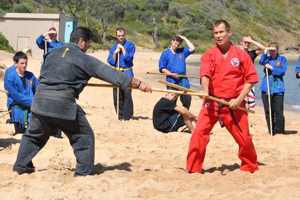 Two men in martial arts gear spar with wooden staffs on a sandy outdoor field, others watching nearby.