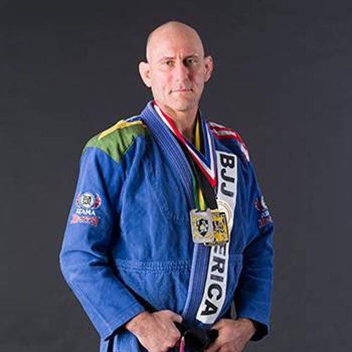 A man in a blue gi with medals and a white-black belt, standing against a gray background.