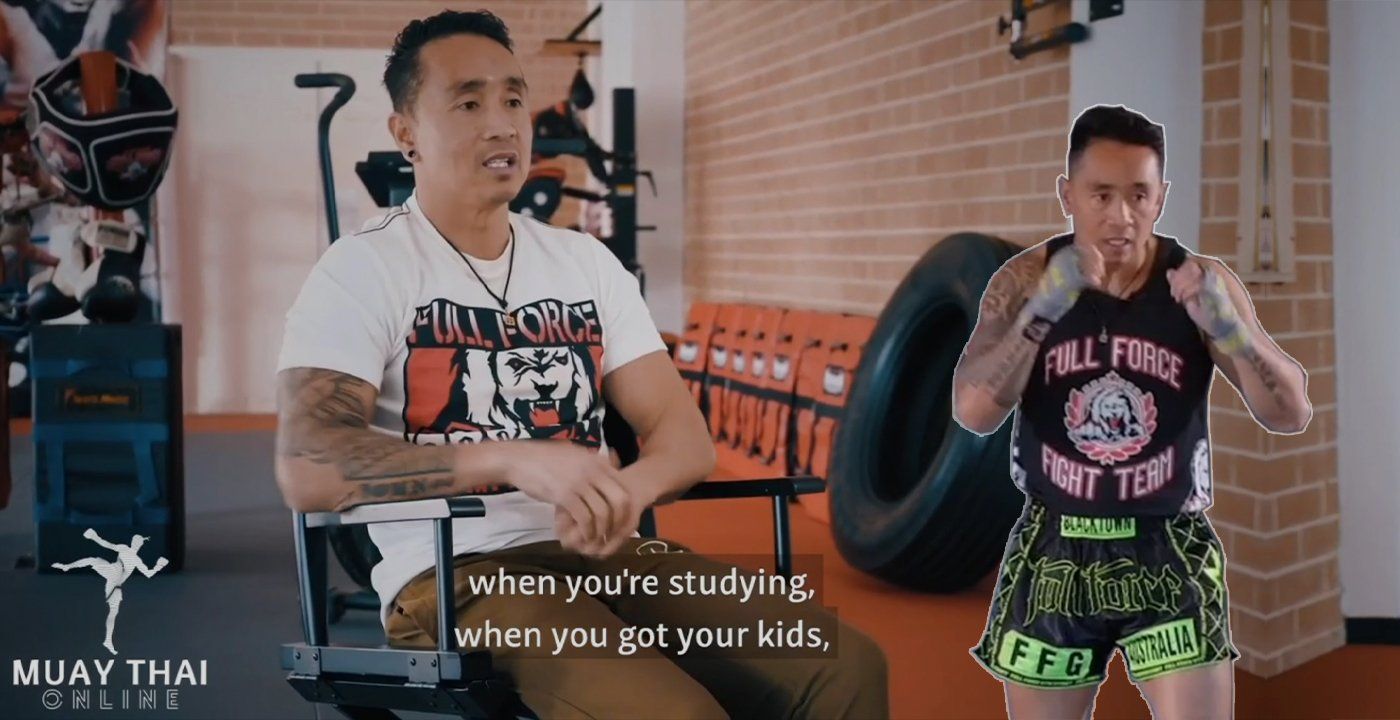 Two men training in a Muay Thai gym, one speaking while the other holds a fighting stance.