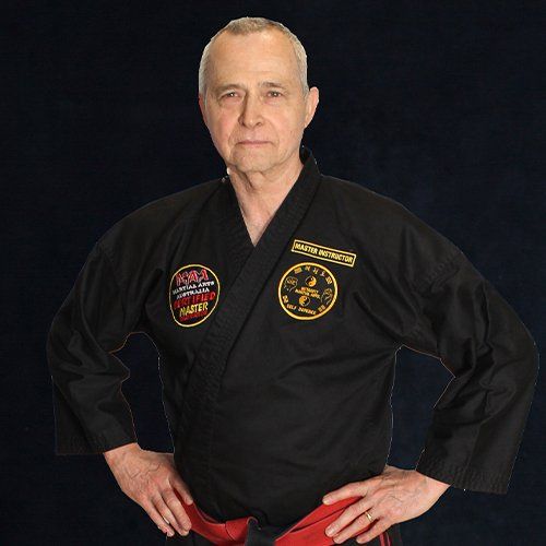 Man in a black martial arts uniform with patches, standing hands on hips against a dark background