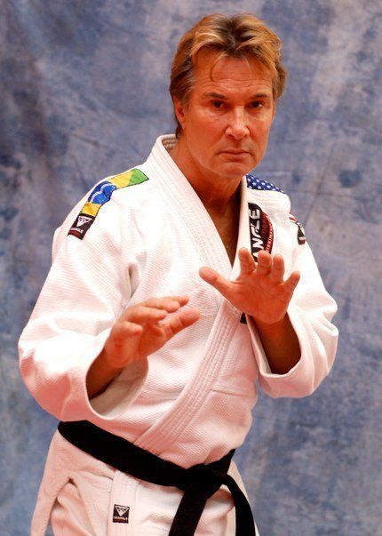 Martial artist in a white gi with black belt, posing in a defensive stance against a blue backdrop