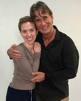 Two smiling people hugging against a plain light-colored wall.