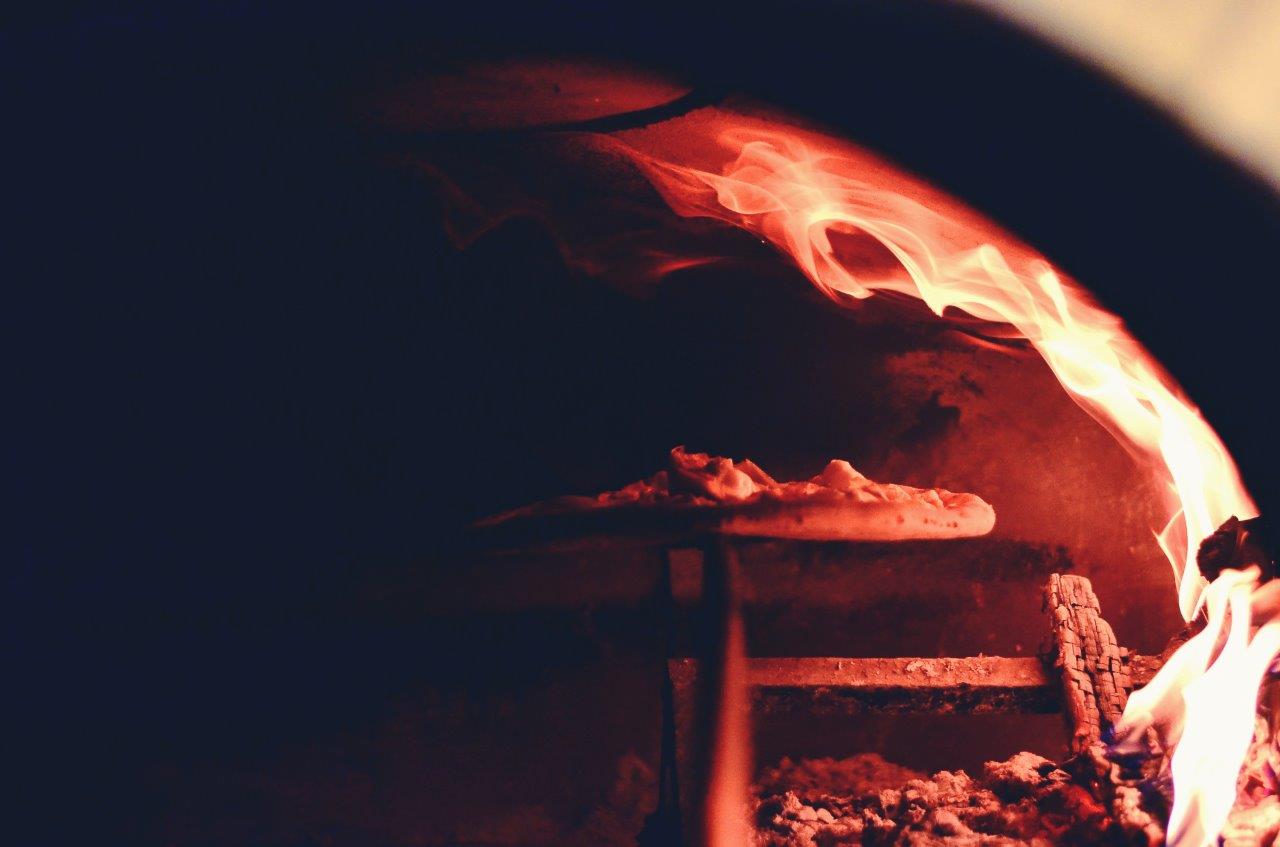 Pizza cotta nel forno a legna con fiamme visibili.
