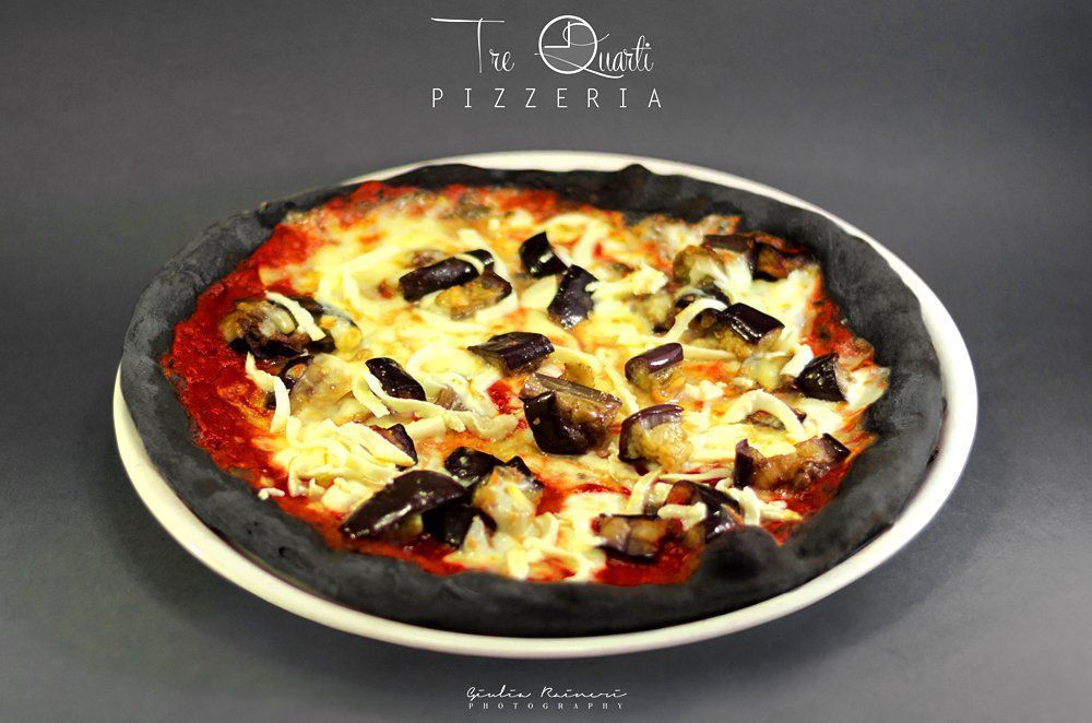 Pizza con crosta nera, guarnita con melanzane e formaggio su un piatto bianco.